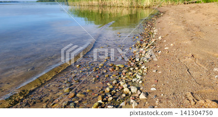 A soft wave rolls over pebbles and clear shallow water toward a sandy shore. Concept of flow, blue mind, grounding, and calm water in motion. A soft wave rolls over pebbles and clear shallow water toward a sandy shore. Concept of flow, blue mind, grounding, and calm water in motion. 141304750