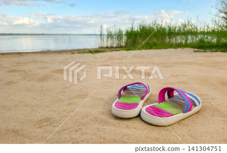 Bright colorful sandals rest in the sand on a lake beach with reeds beyond. Concept of summer vacation, leisure, and beach lifestyle with copy space. 141304751