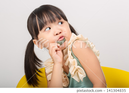 A cute little girl with pigtails sitting on a yellow chair and looking sideways while enjoying a crispy seaweed snack. A cute little girl with pigtails sitting on a yellow chair and looking sideways while enjoying a crispy seaweed snack. 141304835