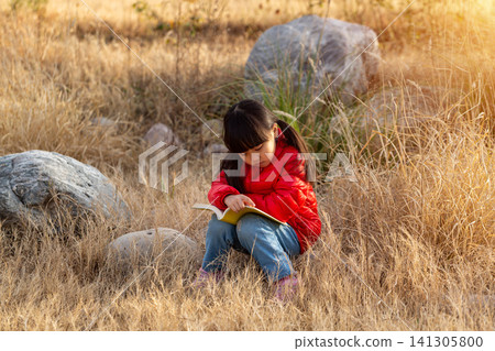 Close up shot of a school girl with a ponytail wearing a red puffer jacket, sitting amongst dry grass and rocks and reading a book, with warm sunset light and blurred background. Close up shot of a school girl with a ponytail wearing a red puffer jacket, sitting amongst dry grass and rocks and reading a book, with warm sunset light and blurred background. 141305800