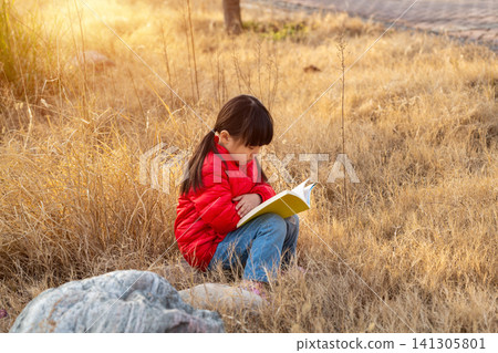 Close up shot of a school girl with a ponytail wearing a red puffer jacket, sitting amongst dry grass and rocks and reading a book, with warm sunset light and blurred background. 141305801