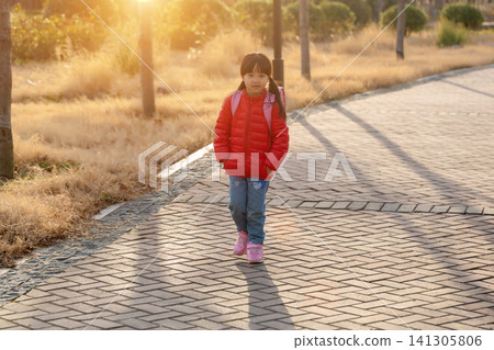 Full shot of a focused young girl with a ponytail wearing a red down jacket and carrying a pink school backpack, walking along a brick paved path in a park at sunset. Full shot of a focused young girl with a ponytail wearing a red down jacket and carrying a pink school backpack, walking along a brick paved path in a park at sunset. 141305806