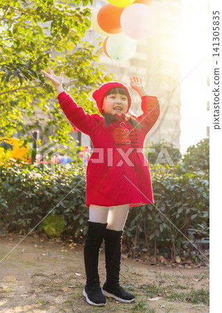 Full vertical shot of a happy school girl with pigtails in a red wool coat and white tights, standing with arms wide open in a sunlit playground with colorful balloons in the background. Full vertical shot of a happy school girl with pigtails in a red wool coat and white tights, standing with arms wide open in a sunlit playground with colorful balloons in the background. 141305835