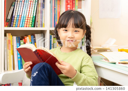 Focused little Asian girl reading a book while sitting on a chair in a home library with bookshelves background. Focused little Asian girl reading a book while sitting on a chair in a home library with bookshelves background. 141305860