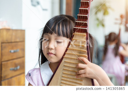 Young Chinese girl practicing Pipa traditional musical instrument on a sofa at home, creative arts and music education. Young Chinese girl practicing Pipa traditional musical instrument on a sofa at home, creative arts and music education. 141305865