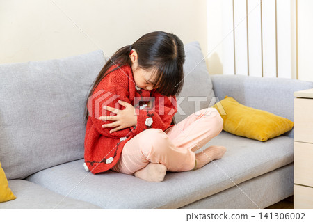 Mid shot of a young girl with pigtails wearing a white shirt and blue denim vest, sitting on a grey sofa and screaming with simulated pain while holding her stomach in a living room. Mid shot of a young girl with pigtails wearing a white shirt and blue denim vest, sitting on a grey sofa and screaming with simulated pain while holding her stomach in a living room. 141306002