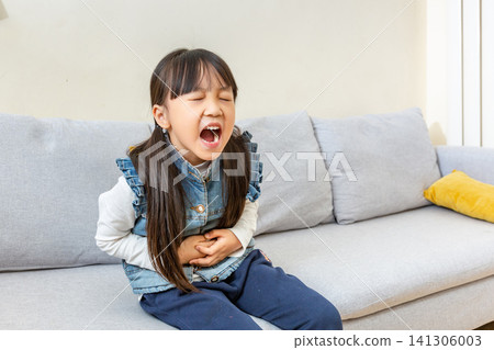 Mid shot of a young girl with pigtails wearing a white shirt and blue denim vest, sitting on a grey sofa and screaming with simulated pain while holding her stomach in a living room. Mid shot of a young girl with pigtails wearing a white shirt and blue denim vest, sitting on a grey sofa and screaming with simulated pain while holding her stomach in a living room. 141306003