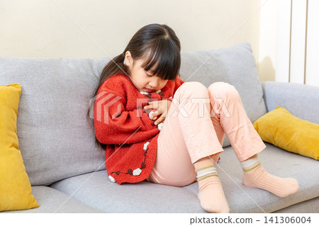 Mid shot of a young girl with pigtails wearing a white shirt and blue denim vest, sitting on a grey sofa and screaming with simulated pain while holding her stomach in a living room. Mid shot of a young girl with pigtails wearing a white shirt and blue denim vest, sitting on a grey sofa and screaming with simulated pain while holding her stomach in a living room. 141306004
