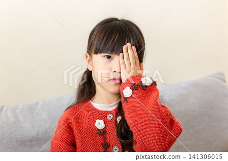 Cute East Asian primary student girl standing alone indoors and covering one eye with her palm, looking at camera 141306015