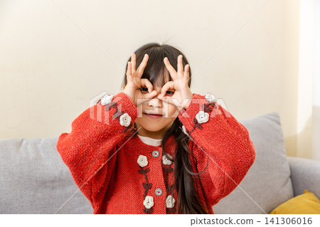 Playful East Asian preschool-age girl standing inside and pretending to form a telescope with both hands making circle gestures near her eyes Playful East Asian preschool-age girl standing inside and pretending to form a telescope with both hands making circle gestures near her eyes 141306016