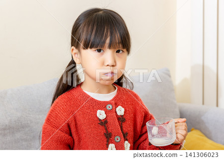 Young East Asian girl with cheeks puffed holding a glass cup with milk and looking at the camera with a full mouth Young East Asian girl with cheeks puffed holding a glass cup with milk and looking at the camera with a full mouth 141306023