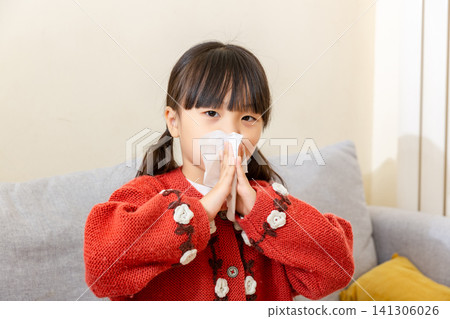Little girl holding a crumpled white tissue against her nose with two hands. 141306026