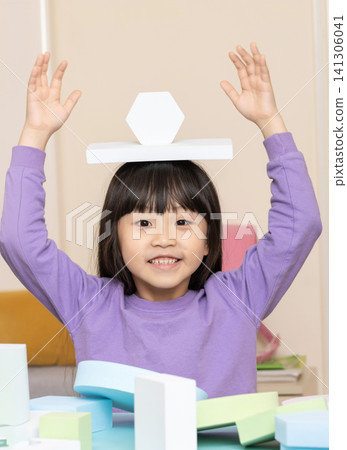 Enthusiastic Asian school-age girl smiling broadly while balancing a hexagonal and a rectangular white foam block on her head, arms raised Enthusiastic Asian school-age girl smiling broadly while balancing a hexagonal and a rectangular white foam block on her head, arms raised 141306041