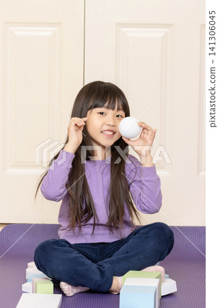 Focused Asian girl sitting cross-legged on a purple yoga mat and holding a white plastic ball near her ear, listening or looking 141306045