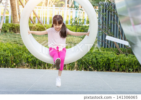 Full shot of a young girl with pigtails sitting alone and swinging happily high on a suspended plastic circle swing at a playground Full shot of a young girl with pigtails sitting alone and swinging happily high on a suspended plastic circle swing at a playground 141306065