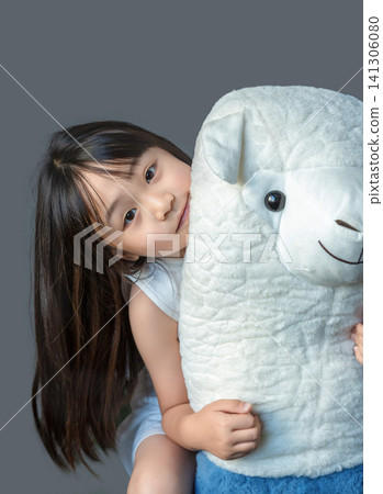 A happy young girl hugging a large white plush alpaca toy against a studio background. 141306080