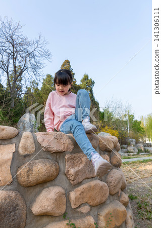Joyful little Asian girl sitting cross-legged by stone wall 141306111