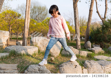 A young girl walking carefully across large rocks in a natural park setting. 141306116