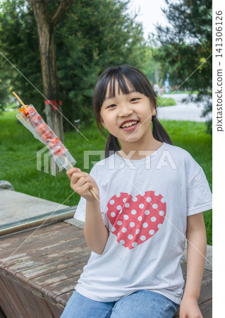 Happy Asian little girl eating tanghulu candied hawthorn skewer outdoors, traditional Chinese street food and joyful childhood concept Happy Asian little girl eating tanghulu candied hawthorn skewer outdoors, traditional Chinese street food and joyful childhood concept 141306126