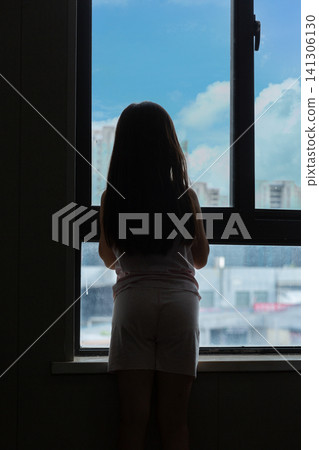 Silhouette of a lonely little girl looking out of the window at city buildings, childhood solitude and contemplation concept. Silhouette of a lonely little girl looking out of the window at city buildings, childhood solitude and contemplation concept. 141306130