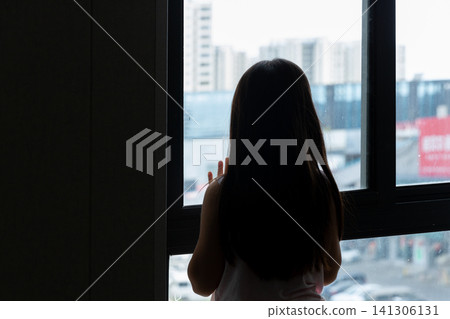 Silhouette of a lonely little girl looking out of the window at city buildings, childhood solitude and contemplation concept. Silhouette of a lonely little girl looking out of the window at city buildings, childhood solitude and contemplation concept. 141306131