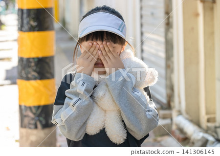 Sad and lonely little girl crying and hugging teddy bear standing by the wall outdoors Sad and lonely little girl crying and hugging teddy bear standing by the wall outdoors 141306145