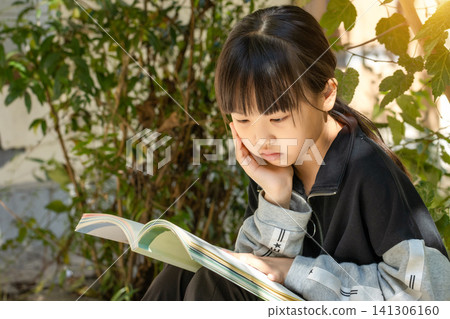 Cute little girl sitting outdoors and reading a book in the garden, concept of nature-based learning and education. Cute little girl sitting outdoors and reading a book in the garden, concept of nature-based learning and education. 141306160