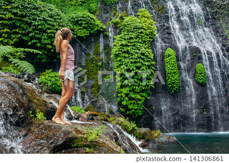Young woman look at view of falling down water of Banyumala Twin Waterfalls. Water flowing under cascading high waterfall. Adventure lifestyle at family holidays travel in Bali tropical rainforest. Young woman look at view of falling down water of Banyumala Twin Waterfalls. Water flowing under cascading high waterfall. Adventure lifestyle at family holidays travel in Bali tropical rainforest. 141306861