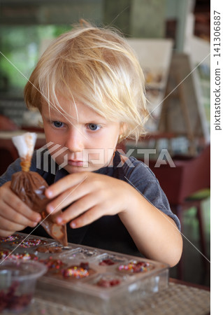 Blond boy carefully squeezes melted chocolate into molds at a workshop in Bali. A hands-on, educational activity for children traveling with family. 141306887