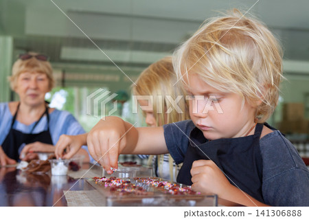 Kids and their mother participate in a chocolate making workshop in Bali, decorating sweets with toppings. A fun and educational family activity during tropical holidays. 141306888