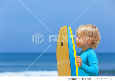 Portrait of little surfer boy with bodyboard have fun on beach, walk by white sand beach. Summer adventures, active families, parents and kids lifestyle, sports activities on school holiday with kids Portrait of little surfer boy with bodyboard have fun on beach, walk by white sand beach. Summer adventures, active families, parents and kids lifestyle, sports activities on school holiday with kids 141306889