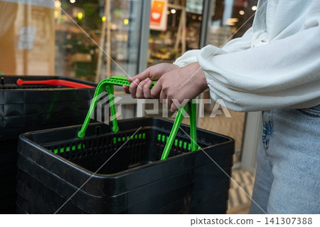 A Close-Up of Hands Grasping the Handle of a Green Shopping Basket Outside a Retail Store with a Modern Aesthetic 141307388