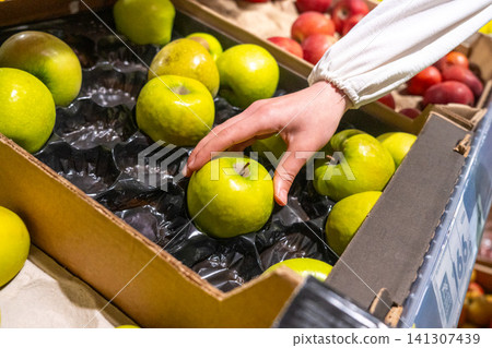 A Person Selecting Fresh Green Apples from a Cardboard Box in a Vibrant Produce Section of a Market. A Person Selecting Fresh Green Apples from a Cardboard Box in a Vibrant Produce Section of a Market. 141307439