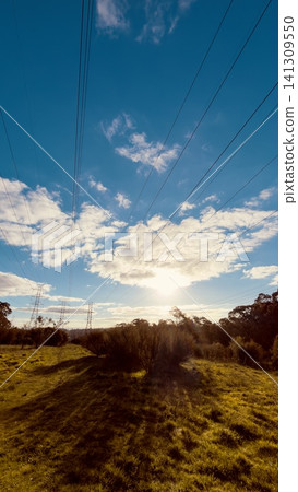 Low angle perspective of high voltage power lines cutting through a grassy countryside landscape against a bright sunset sky Low angle perspective of high voltage power lines cutting through a grassy countryside landscape against a bright sunset sky 141309550