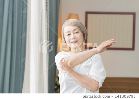 A senior woman doing morning stretches by the living room window. A senior woman doing morning stretches by the living room window. 141309795