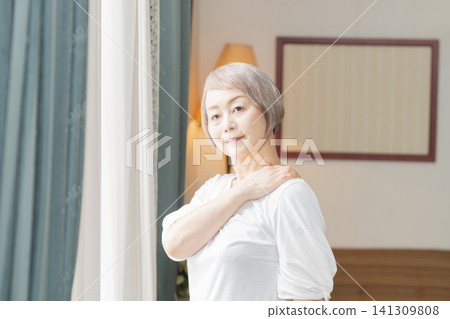 一位老婦人在客廳窗邊做晨間伸展運動。 一位老婦人在客廳窗邊做晨間伸展運動。 141309808