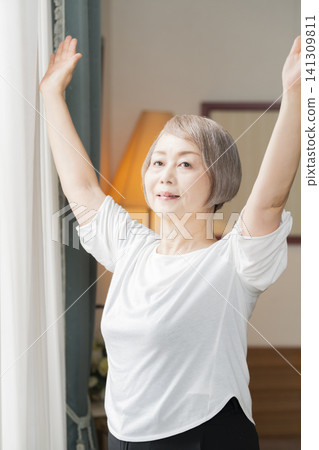 A senior woman doing morning stretches by the living room window. A senior woman doing morning stretches by the living room window. 141309811