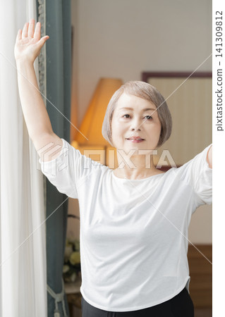 一位老婦人在客廳窗邊做晨間伸展運動。 一位老婦人在客廳窗邊做晨間伸展運動。 141309812