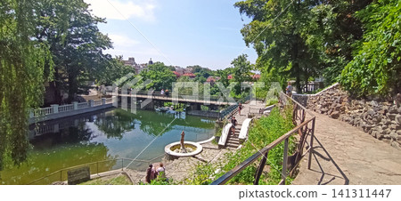 Beautiful park with a calm pond and footbridge in Kamianets-Podilskyi Beautiful park with a calm pond and footbridge in Kamianets-Podilskyi 141311447