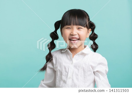A happy East Asian schoolgirl laughing joyfully while standing against a solid mint green studio background. A happy East Asian schoolgirl laughing joyfully while standing against a solid mint green studio background. 141311503