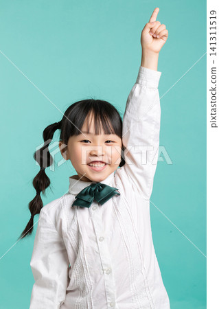 A cheerful young schoolgirl raising her hand high to answer a question during a classroom activity in a studio setting. A cheerful young schoolgirl raising her hand high to answer a question during a classroom activity in a studio setting. 141311519