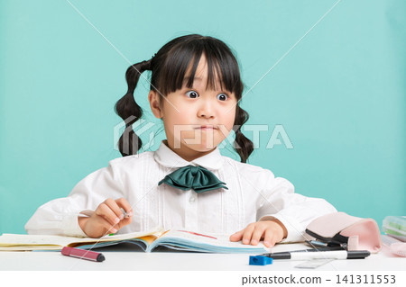 A surprised young girl sitting at a white school desk with stationery supplies, looking up with wide eyes during her study. A surprised young girl sitting at a white school desk with stationery supplies, looking up with wide eyes during her study. 141311553