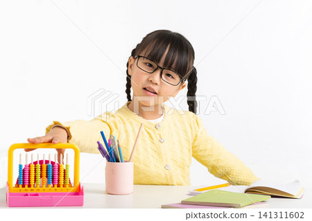 Focused little Asian girl using a colorful abacus on a white desk with glasses and pencils. 141311602
