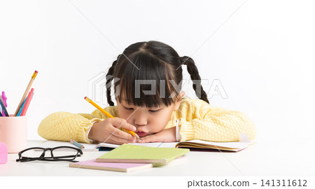 Diligent little Asian girl leaning over and focused on writing with a pencil in a notebook on a white desk. Diligent little Asian girl leaning over and focused on writing with a pencil in a notebook on a white desk. 141311612