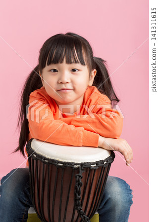 Cute little girl sitting behind a djembe drum with her arms crossed on pink background. Cute little girl sitting behind a djembe drum with her arms crossed on pink background. 141311615