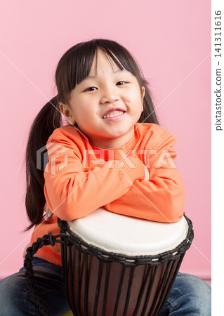Cute little girl sitting behind a djembe drum with her arms crossed on pink background. Cute little girl sitting behind a djembe drum with her arms crossed on pink background. 141311616