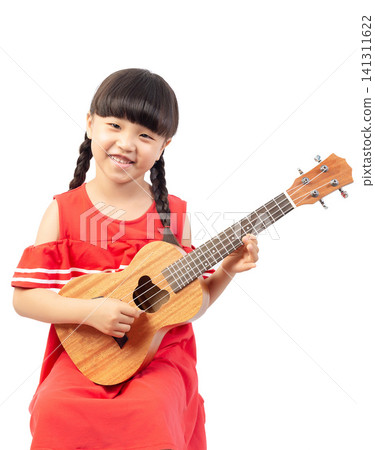 Focused little girl with braids looking down while playing a wooden ukulele against a white background. Focused little girl with braids looking down while playing a wooden ukulele against a white background. 141311622