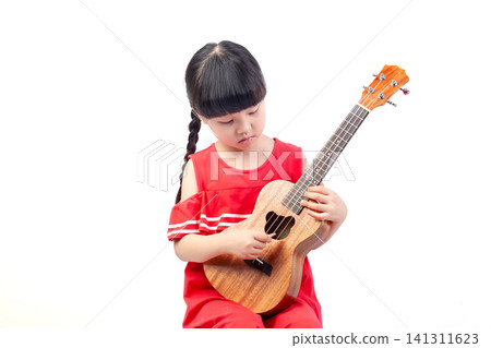 Focused little girl with braids looking down while playing a wooden ukulele against a white background. Focused little girl with braids looking down while playing a wooden ukulele against a white background. 141311623