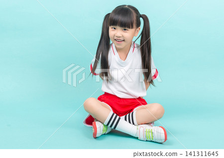 Happy little girl in sportswear sitting cross-legged on the floor against a solid light blue backdrop. Happy little girl in sportswear sitting cross-legged on the floor against a solid light blue backdrop. 141311645