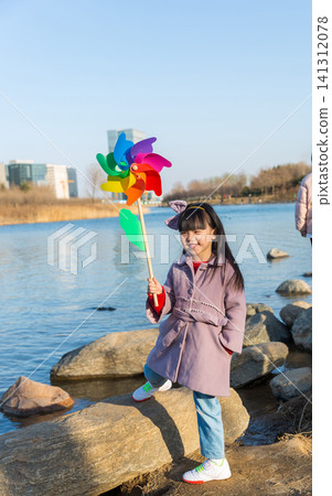 a happy little girl stands on a riverside rock, holding a colorful pinwheel. a happy little girl stands on a riverside rock, holding a colorful pinwheel. 141312078